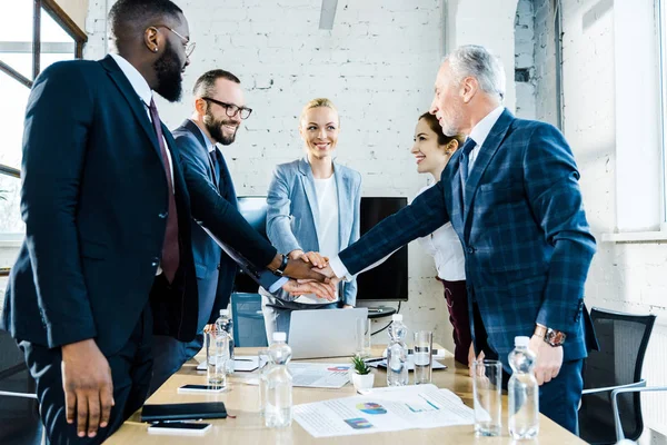 Empresarias alegres poniendo manos juntas con compañeros de trabajo multiculturales - foto de stock