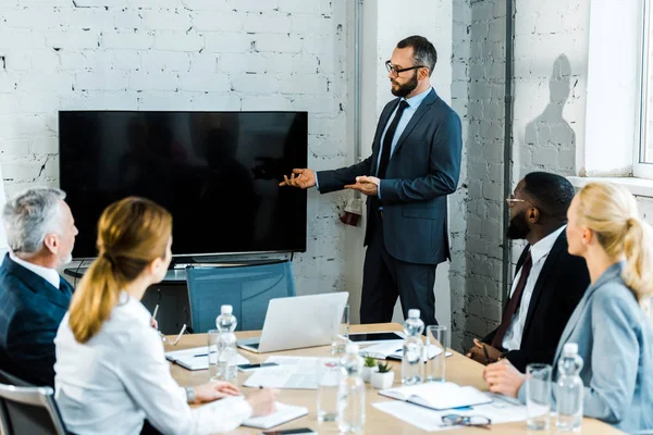 Business coach barbuto in occhiali da vista gesticolando vicino alla tv con schermo bianco e colleghi multiculturali — Foto stock