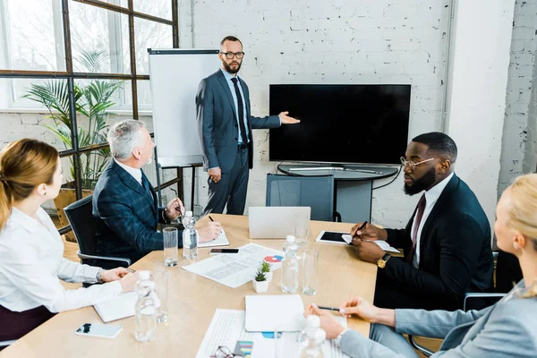 Business coach in formal wear gesturing near tv with blank screen and multicultural coworkers — Stock Photo