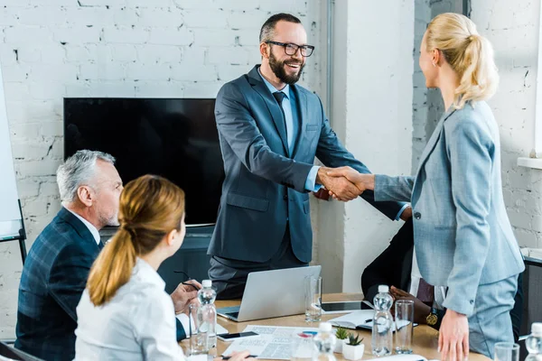 Apuesto entrenador de negocios estrechando la mano con la mujer rubia cerca de compañeros de trabajo multiculturales - foto de stock