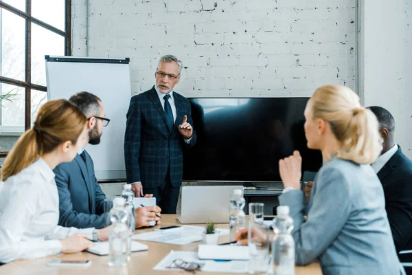 Selektiver Fokus des Businesstrainers gestikuliert in der Nähe des Fernsehers mit leerem Bildschirm und multikulturellen Menschen — Stockfoto