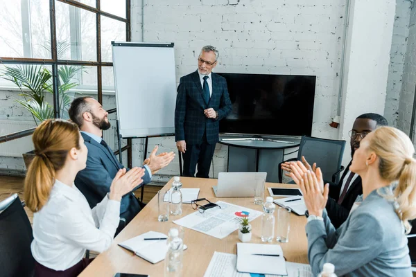 Enfoque selectivo del entrenador de negocios de pie cerca de la pizarra blanca cerca de compañeros de trabajo multiculturales aplaudiendo de la mano en la sala de conferencias - foto de stock