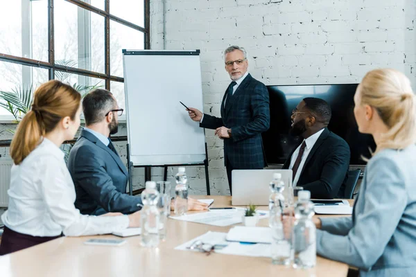 Selective focus of business coach pointing at white board with pen near multicultural coworkers — Stock Photo