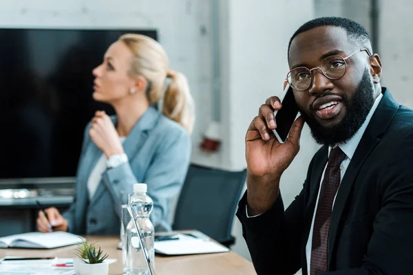 Enfoque selectivo de hombre afroamericano hablando en el teléfono inteligente cerca de compañero de trabajo rubia - foto de stock