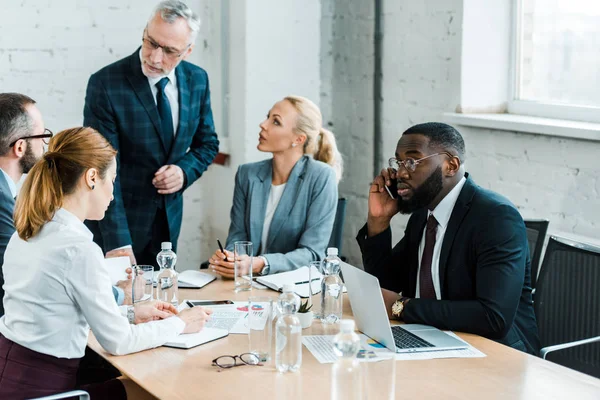 Selektiver Fokus eines afrikanisch-amerikanischen Mannes, der in der Nähe von Laptop und Kollegen mit dem Smartphone spricht — Stockfoto