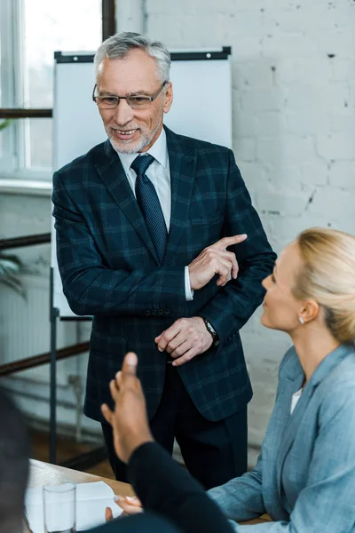 Selective focus of cheerful business coach in glasses pointing with finger near multicultural coworkers — Stock Photo