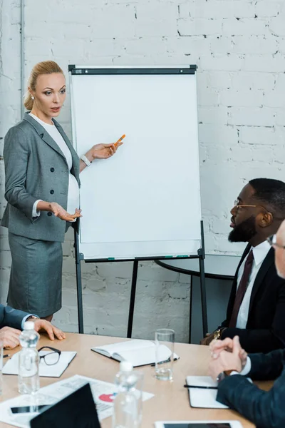 Selective focus of attractive pregnant business coach standing near white board and gesturing in conference room — Stock Photo