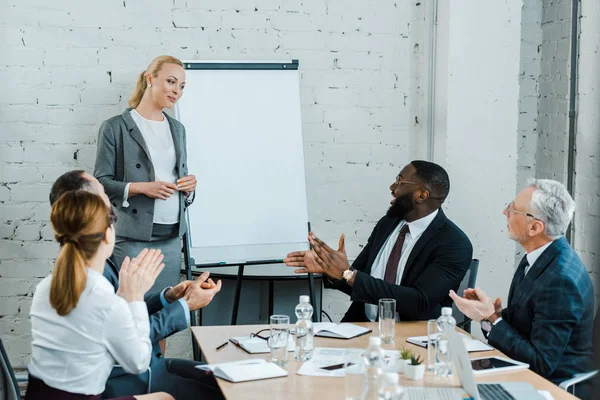 Personas multiculturales aplaudiendo al entrenador de negocios embarazada de pie cerca de la pizarra blanca - foto de stock