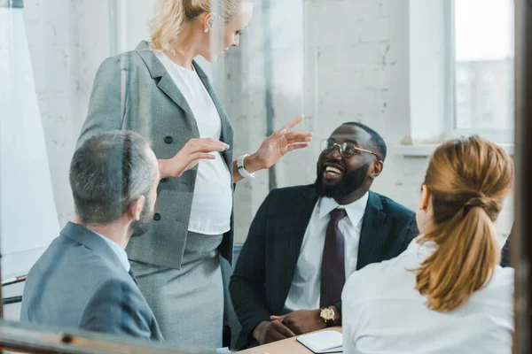 Atractivo rubia embarazada negocio entrenador gesto mientras de pie cerca de africano americano hombre - foto de stock