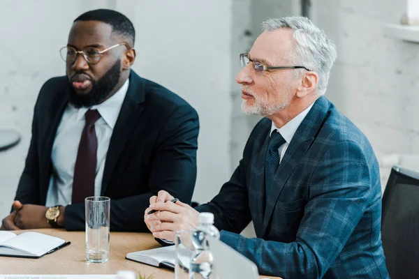 Bearded businessman in eye glasses sitting near african american coworker in conference room — Stock Photo