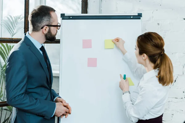 Geschäftsfrau legt klebrige Zettel auf Whiteboard, während sie mit Geschäftsmann in Brille steht — Stockfoto