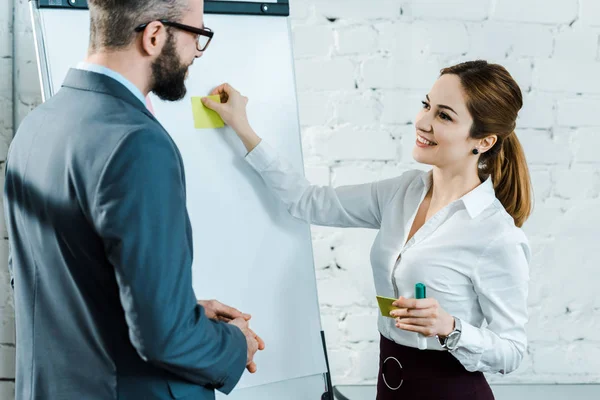 Fröhliche Geschäftsfrau legt klebrige Zettel auf weiße Tafel, während sie mit Geschäftsmann in Brille steht — Stockfoto