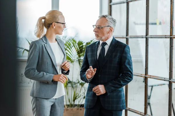 Atractiva mujer de negocios rubia haciendo gestos mientras habla con un hombre de negocios barbudo en gafas para los ojos - foto de stock