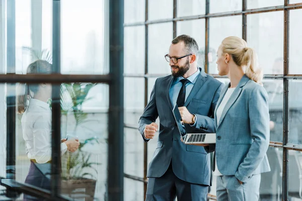 Веселий бородатий бізнесмен дивиться на колегу, стоячи поруч з бізнес-леді з ноутбуком — стокове фото