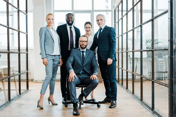 Guapo barbudo hombre de negocios sentado en silla cerca de compañeros de trabajo multiculturales - foto de stock