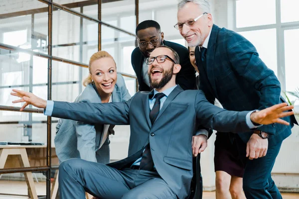 Fröhlicher Geschäftsmann, der mit ausgestreckten Händen neben glücklichen multikulturellen Mitarbeitern auf einem Stuhl sitzt — Stockfoto