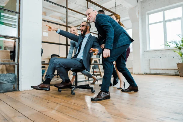 Low angle view of cheerful businessman sitting on chair and pointing with finger near multicultural coworkers — Stock Photo