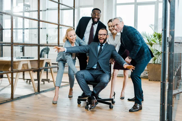 Fröhlicher Geschäftsmann, der mit ausgestreckten Händen neben multikulturellen Mitarbeitern auf einem Stuhl sitzt — Stockfoto