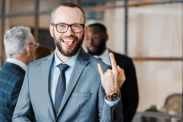 Selective focus of happy businessman showing tongue and rock sign near multicultural coworkers — Stock Photo