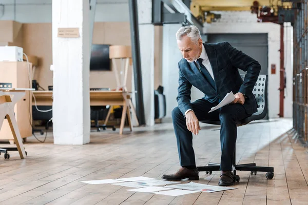 Красивий бородатий бізнесмен сидить на стільці і дивиться на графіки і графіки на підлозі — стокове фото