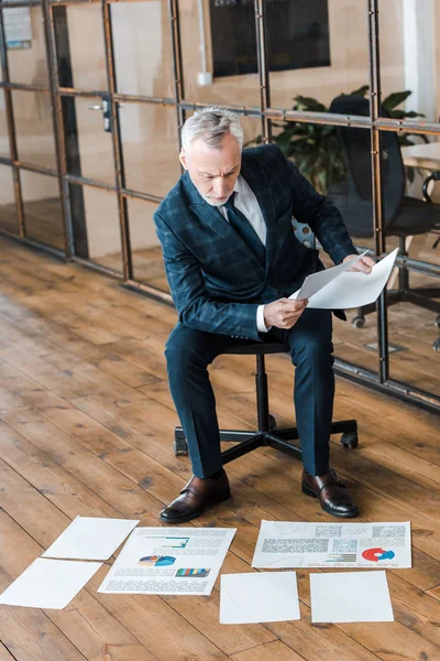 Schöner Geschäftsmann sitzt auf Stuhl und hält Papiere in der Nähe von Diagrammen und Diagrammen — Stockfoto