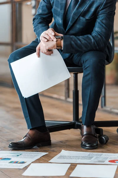 Vista recortada del hombre de negocios sentado en la silla y sosteniendo vacío en blanco cerca de gráficos y gráficos - foto de stock