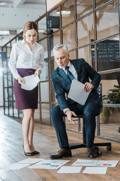 Schöner Mann, der mit dem Finger auf Diagramme und Grafiken zeigt, während er auf einem Stuhl neben Geschäftsfrau sitzt — Stockfoto