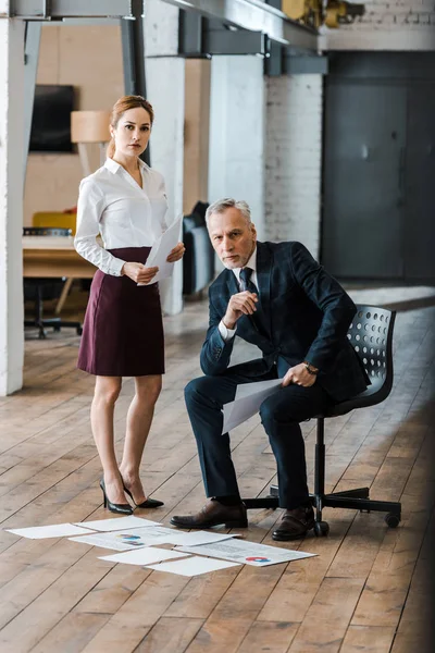 Handsome man sitting on chair near businesswoman and charts and graphs on floor — Stock Photo