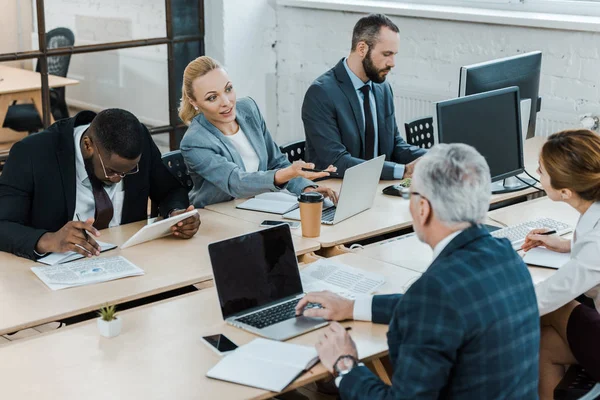 Atractiva mujer de negocios gesto mientras habla con compañero de trabajo cerca de hombre afroamericano - foto de stock