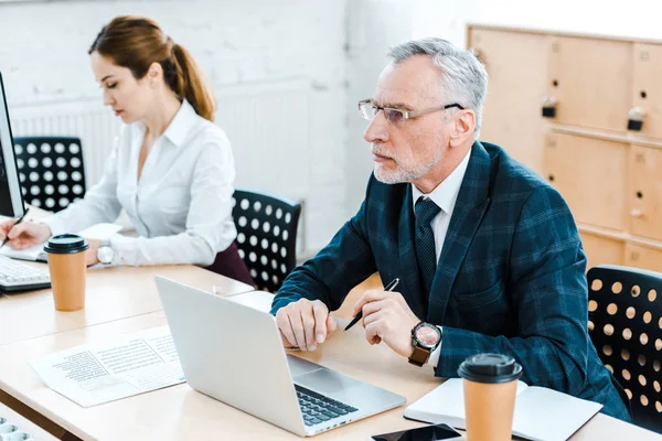 Selective focus of bearded businessman in glasses using laptop near businesswoman in office — Stock Photo