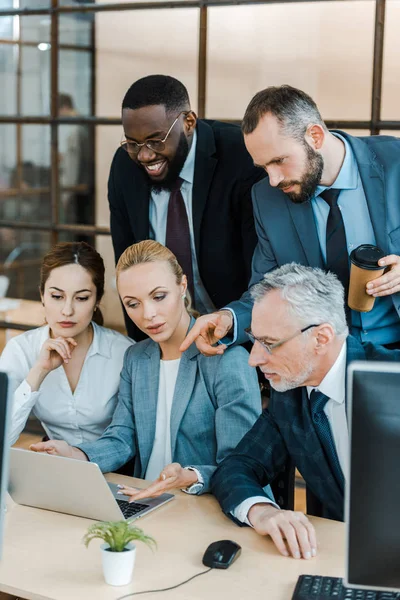 Hombre de negocios barbudo con taza de papel apuntando con el dedo a la computadora portátil cerca de compañeros de trabajo multiculturales - foto de stock