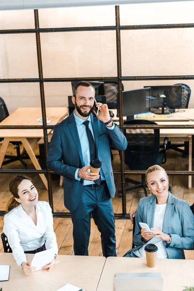 Overhead view of happy bearded businessman talking on smartphone near attractive coworkers — Stock Photo