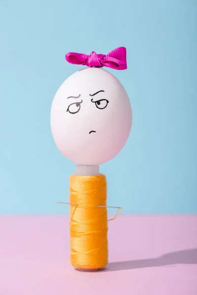 Egg with bow and angry face expression on thread coil on blue and pink — Stock Photo