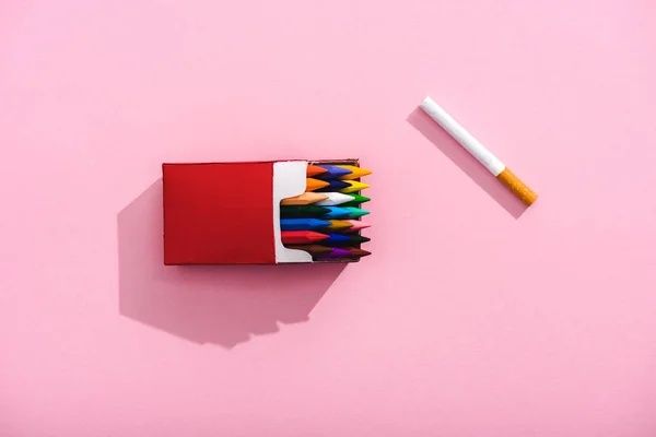 Vue du dessus des crayons colorés dans le paquet de cigarettes et la cigarette de tabac sur rose, concept de tabagisme — Photo de stock