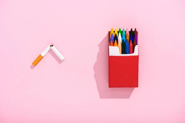 Vue du dessus des crayons colorés dans le paquet de cigarettes et la cigarette de tabac cassée sur rose, concept de tabagisme — Photo de stock