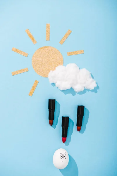 Top view of paper sun, cotton candy cloud, egg with surprised face expression and Lipstick raindrops on blue — Stock Photo