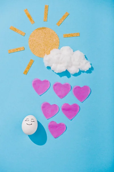 Vista superior de sol de papel, nube de algodón de azúcar, huevo con expresión de cara feliz y gotas de lluvia en forma de corazón en azul - foto de stock