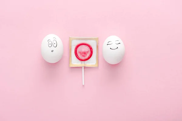 Top view of eggs with happy and surprised face expressions with condom wrapped as lollipop on pink with copy space — Stock Photo