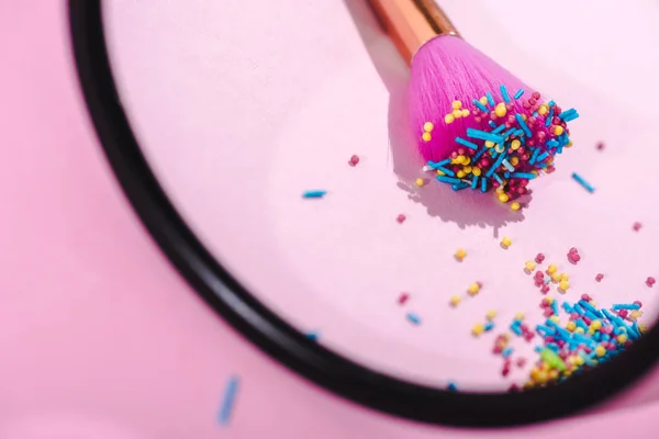 Selective focus of makeup brush covered in colorful sprinkles reflecting in mirror on pink — Stock Photo