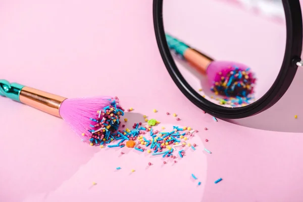 Pincel de maquillaje cubierto de coloridos espolvoreos con reflejo en espejo sobre rosa - foto de stock