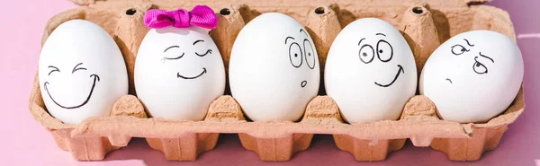 Панорамный снимок яиц с различными выражениями лица в коробке из-под яиц на розовом — стоковое фото