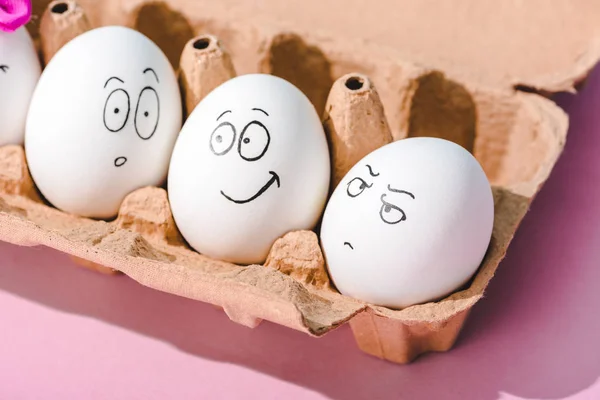 Huevos con expresiones de cara sorprendidas, enojadas y sonrientes en cartón de huevo en rosa - foto de stock