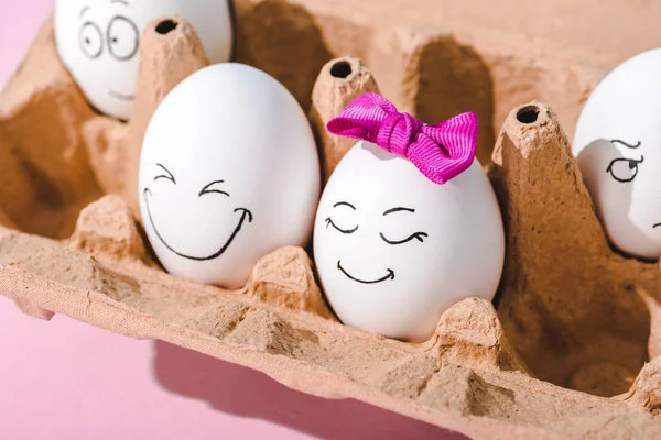 Яйца с различными выражениями лица в коробке с яйцами на розовом — стоковое фото