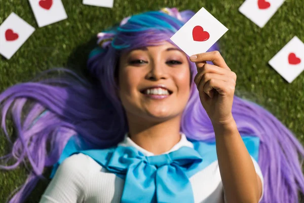 Selective focus of asian anime girl in purple wig holding card and lying on grass — Stock Photo