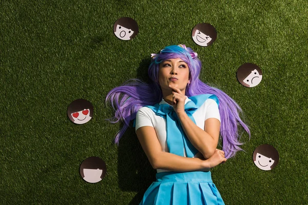 Pensive asian anime girl in purple wig with emoticons lying on grass — Stock Photo