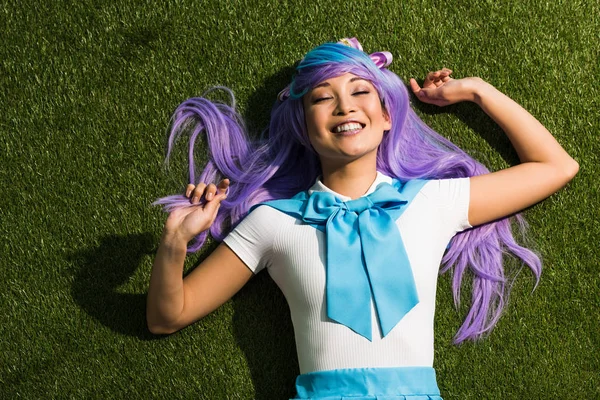 Laughing anime girl in purple wig lying on grass with closed eyes — Stock Photo