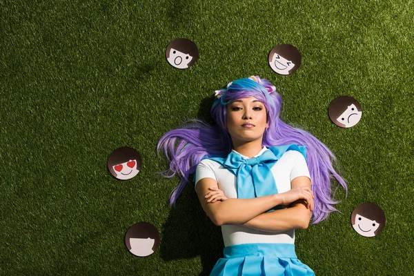 Chica anime asiático en peluca púrpura con emoticonos tumbados en la hierba - foto de stock