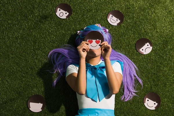 Chica de anime en peluca púrpura acostado en la hierba con emoticonos - foto de stock