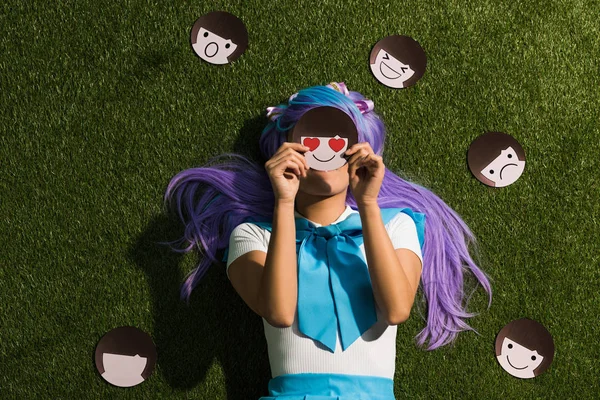 Anime girl in purple wig lying on grass with emoticons — Stock Photo
