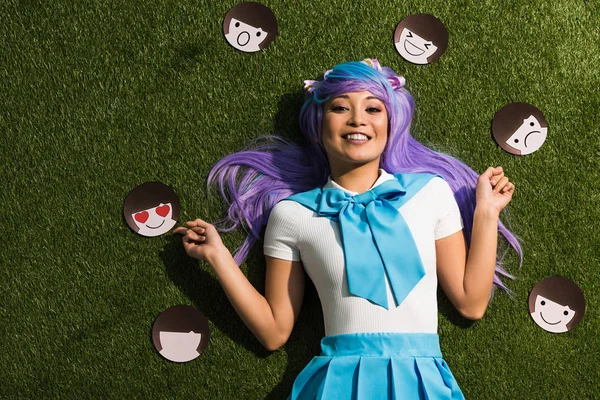 Chica anime asiático en peluca púrpura con emoticonos tumbados en la hierba - foto de stock